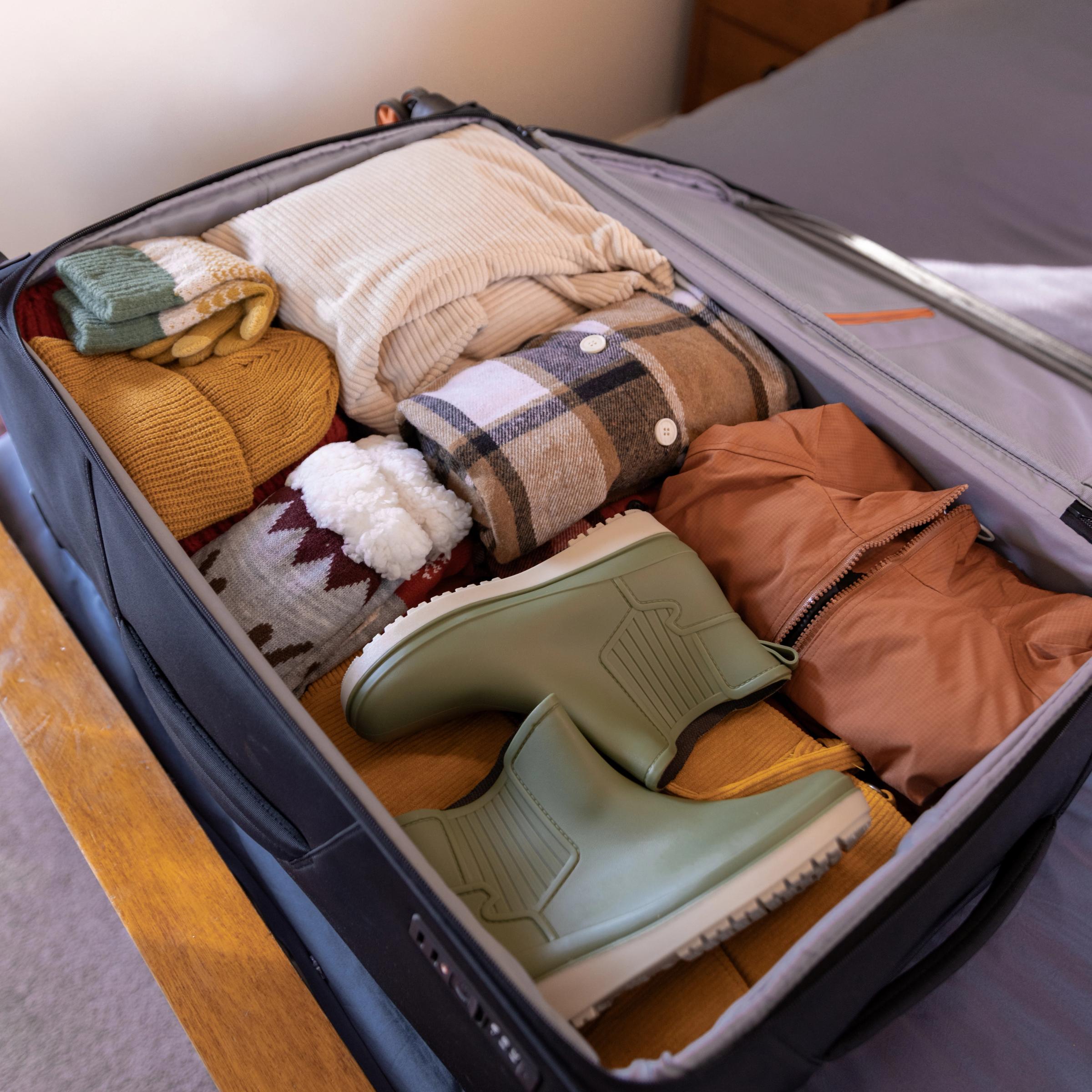 An open suitcase filled with neatly packed warm-toned clothing and sage green rain boots.
