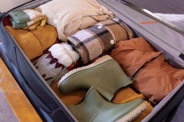 An open suitcase neatly packed with warm clothes and green rain boots.