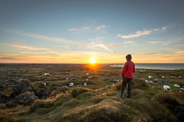 Person on a grass-covered hill overlooking a coastal campsite at sunset.