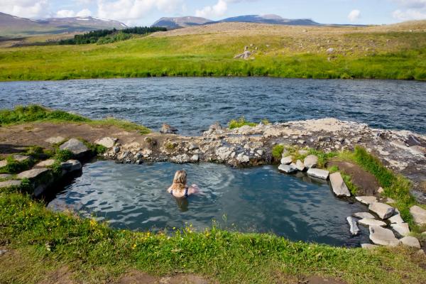 A woman in a natural hot spring next to a river, with green hills and distant mountains.