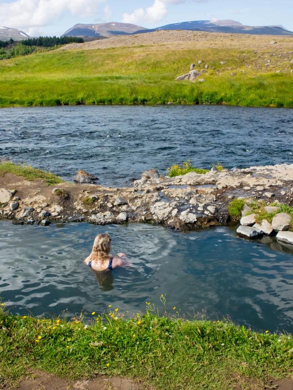A person relaxing in a natural hot spring next to a river with green hills and mountains in the background.
