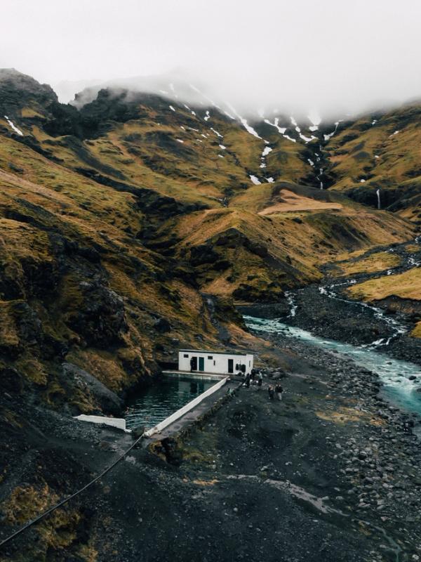 White building with a pool next to a turquoise river in a steep, grassy mountain valley with cloudy peaks.
