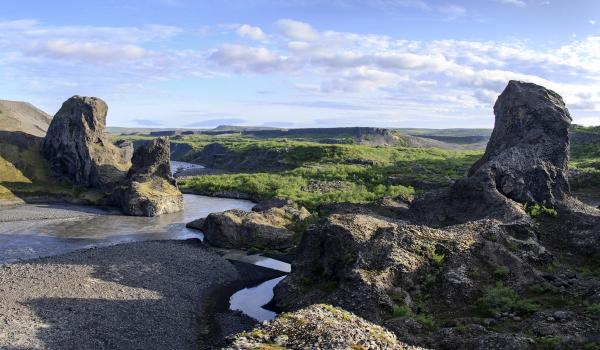 A rugged river landscape with dark rock formations and green vegetation under a partly cloudy sky.