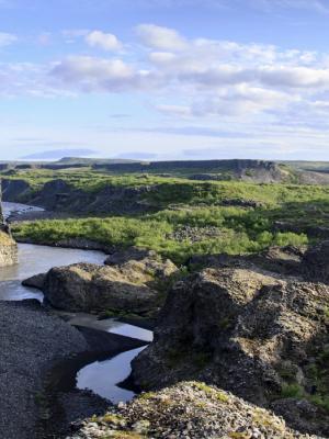 A rugged river landscape with dark rock formations and green vegetation under a partly cloudy sky.