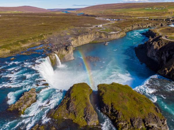 A wide waterfall plunges into a turquoise river with a rainbow arching through its mist, surrounded by a vast, green-brown landscape.