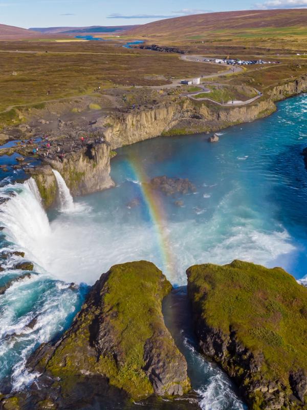 A powerful waterfall with bright blue water and a rainbow arching through its mist, surrounded by rocky, green-brown land.