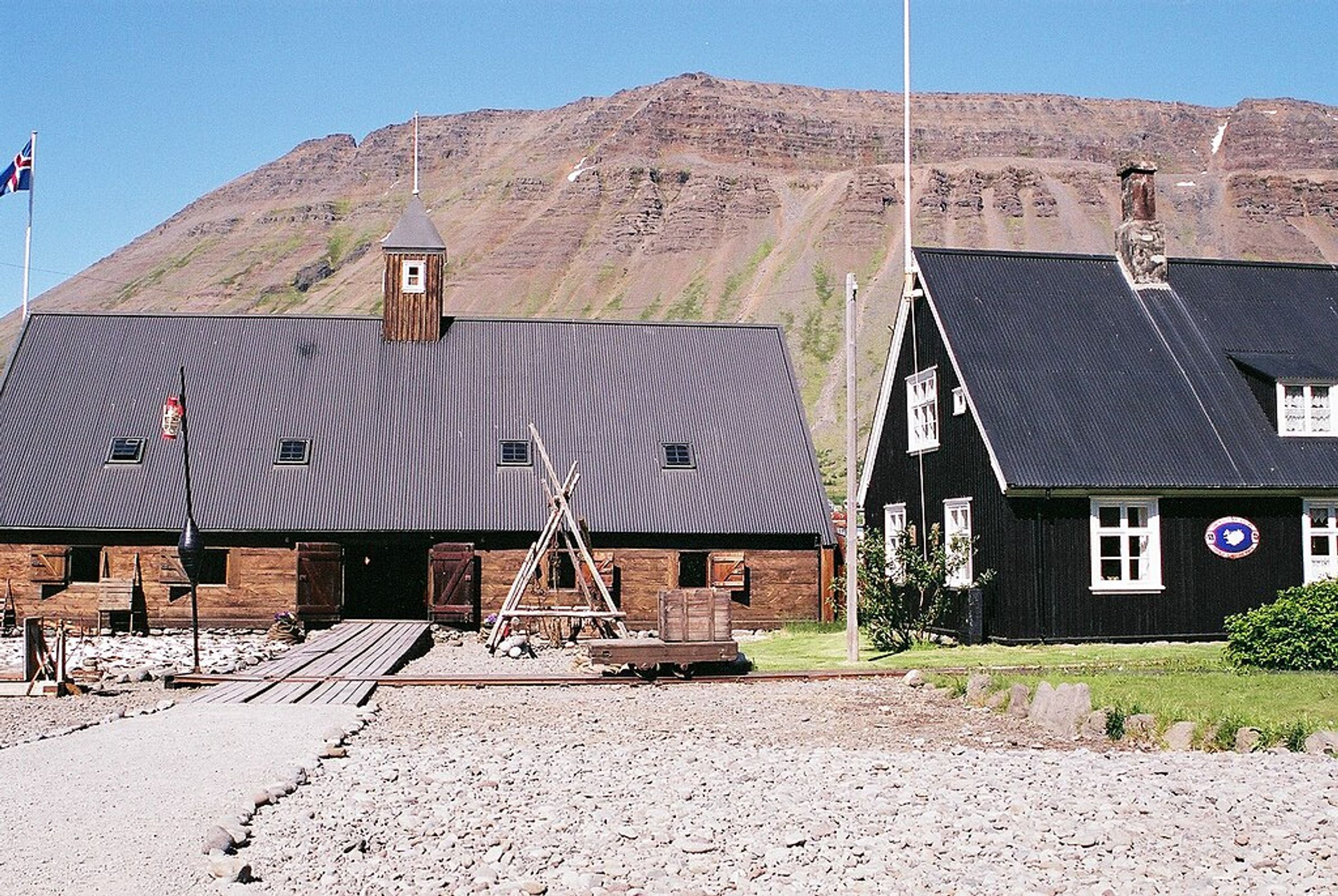 Two buildings, one wooden and one black, stand before a large brown mountain under a blue sky, with an Icelandic flag flying.