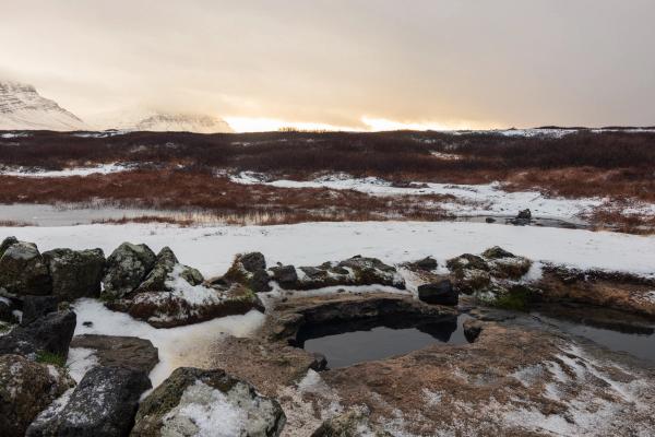 Natural hot spring surrounded by snow in a mountainous winter landscape with a golden sky.