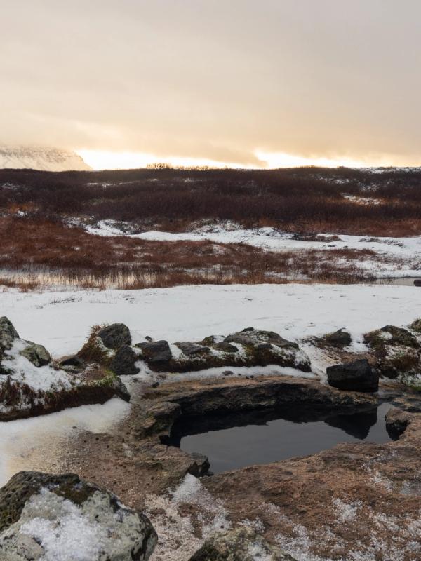 Natural hot spring pool in a snowy, rocky landscape with distant mountains under an orange sky.
