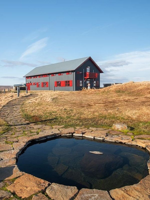 A stone-lined hot spring in the foreground, with a path leading to a dark grey building with red shutters on a grassy hill under a blue sky.