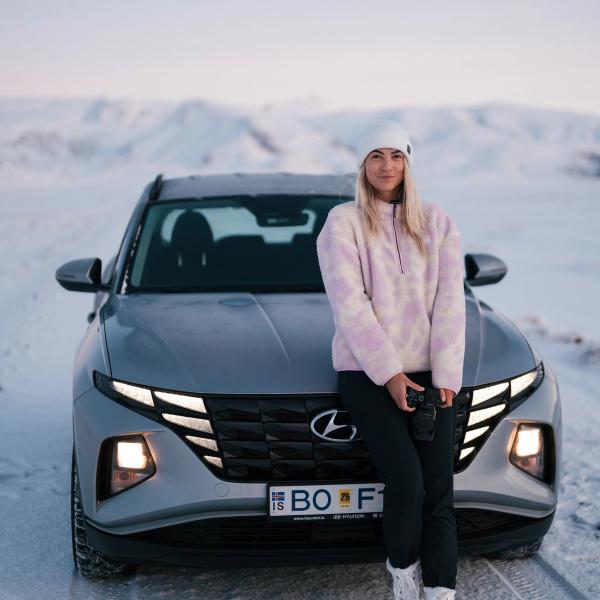 A black Jeep Wrangler with headlights on drives on a snow-covered road, with a red house in the snowy background.