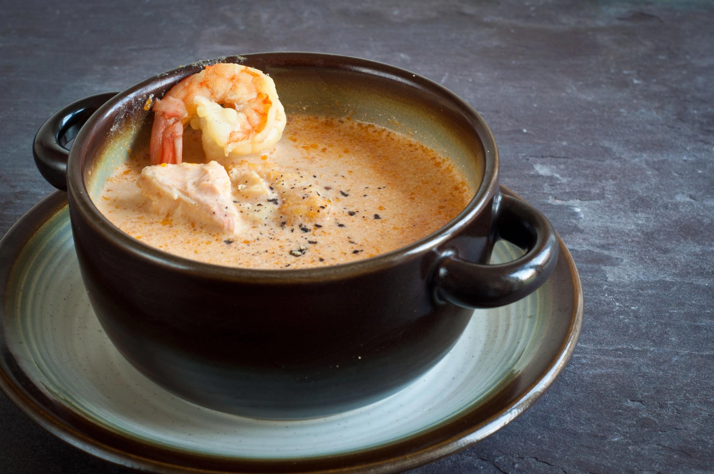 Creamy orange soup with shrimp and fish chunks in a dark brown bowl on a dark surface.
