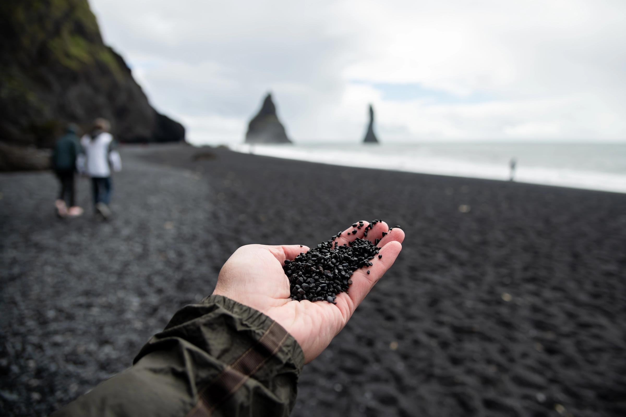 Hand holding black pebbles on a black sand beach with sea stacks in the distance.