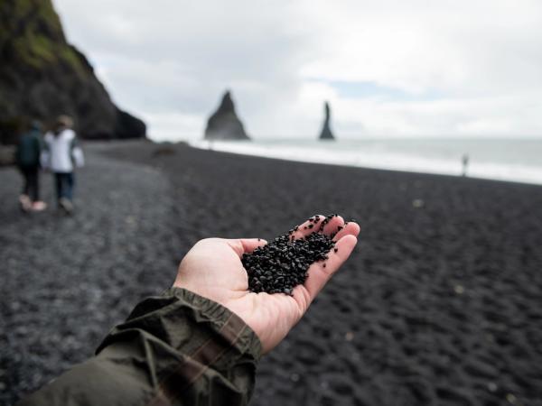 Mano sosteniendo piedras negras en una playa de arena negra con formaciones rocosas en el mar a lo lejos.