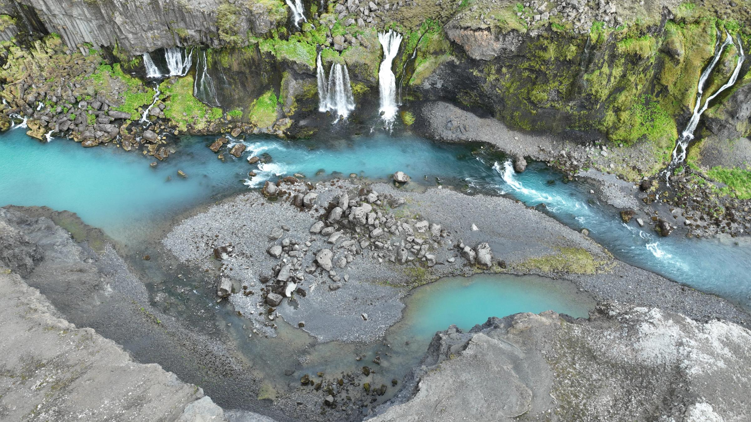 Vista aérea de un río turquesa con múltiples cascadas que caen de un acantilado verde, y una pequeña piscina turquesa.