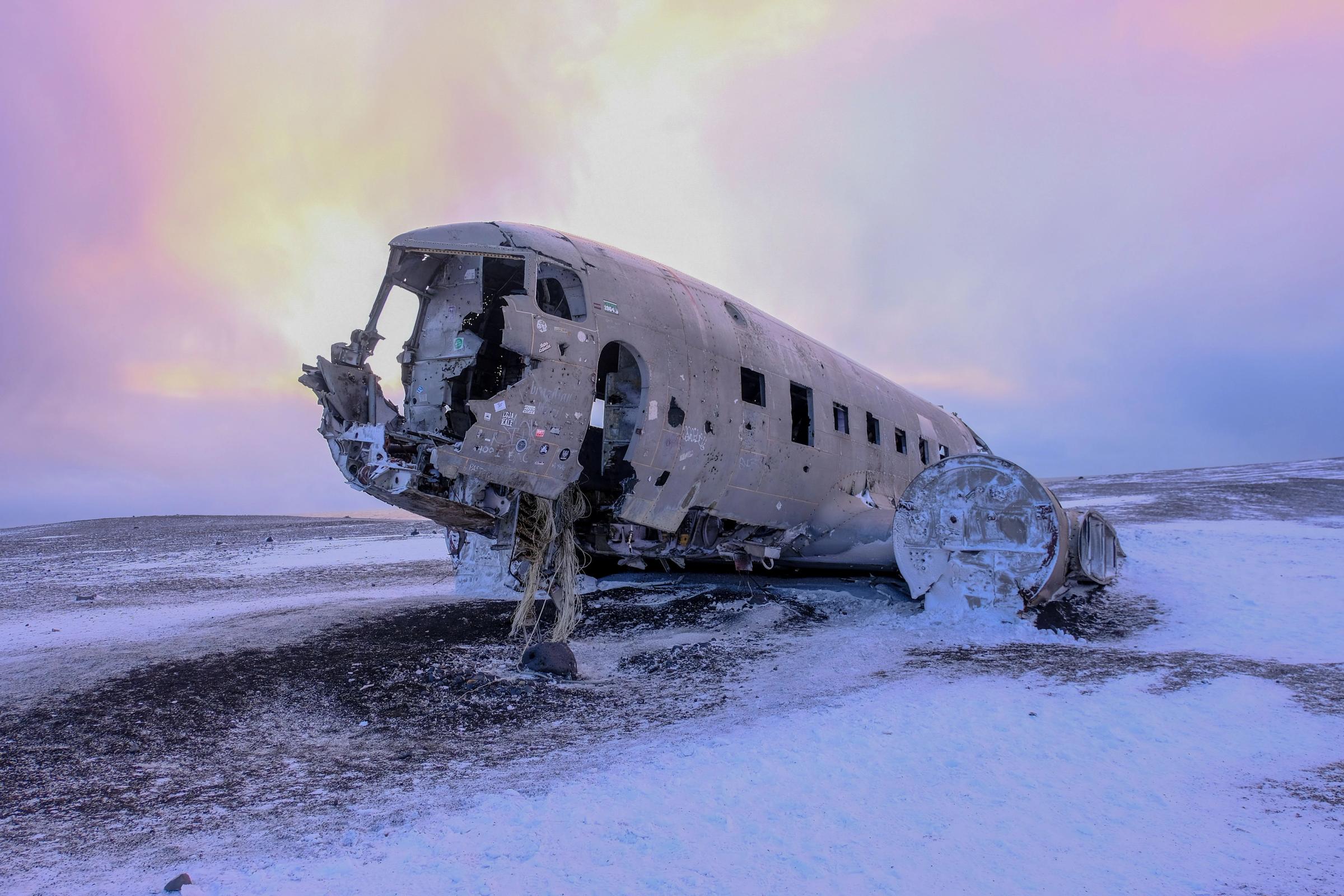 The corroded fuselage of a crashed airplane lies on a snowy, desolate landscape under a dramatic purple and yellow sky.