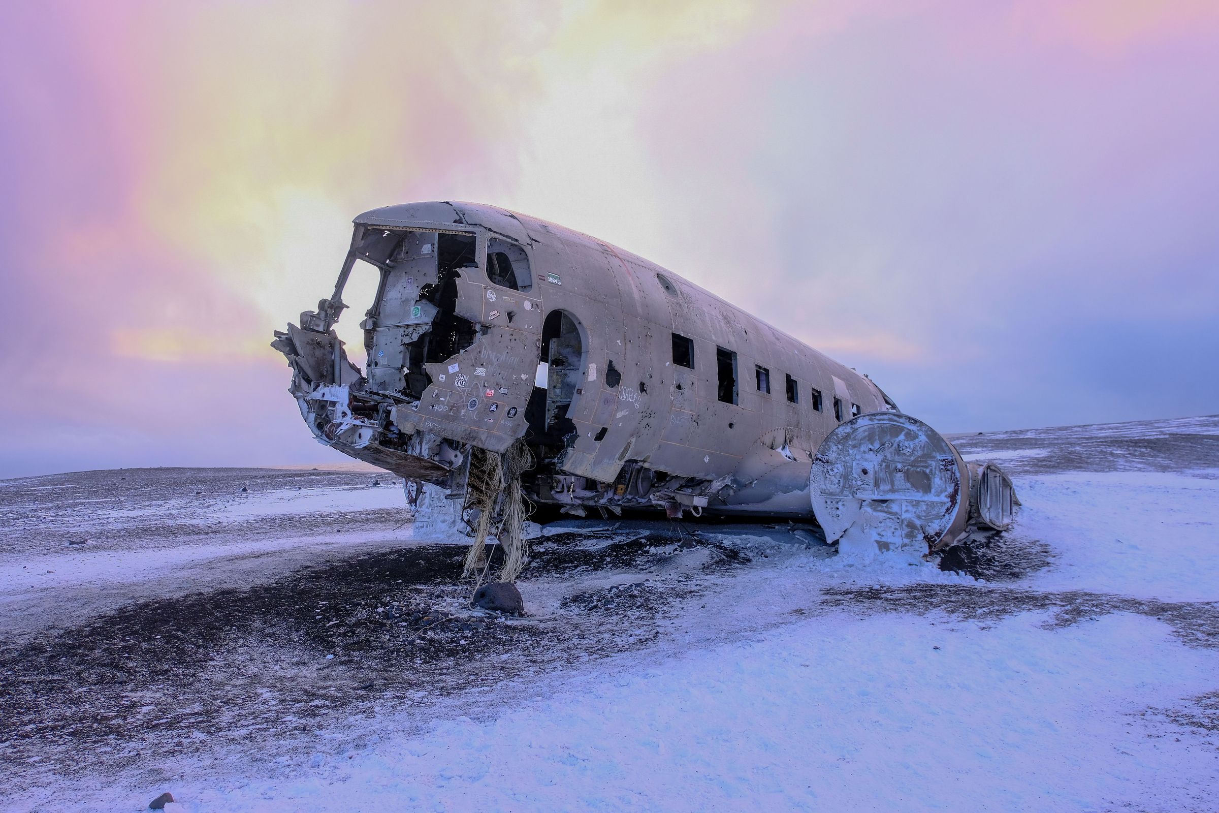 The corroded fuselage of a crashed airplane lies on a snowy, desolate landscape under a dramatic purple and yellow sky.