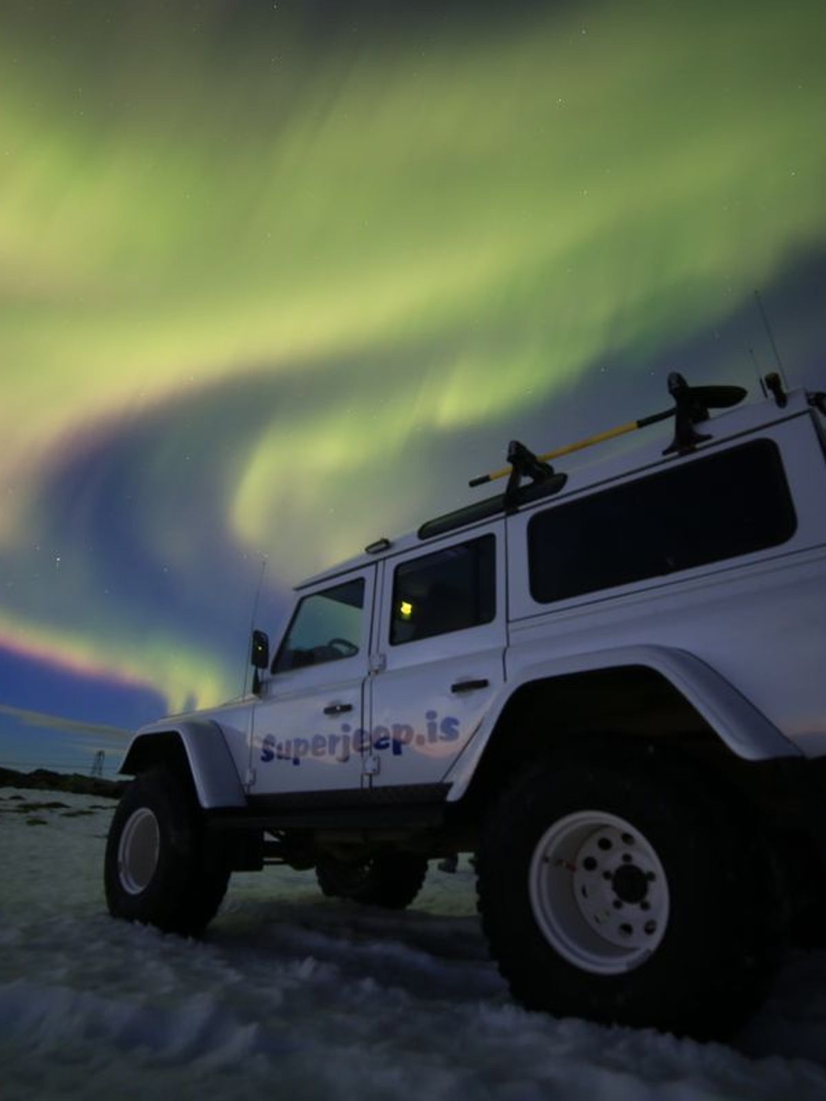 Un jeep blanco aparcado en la nieve bajo la aurora boreal.