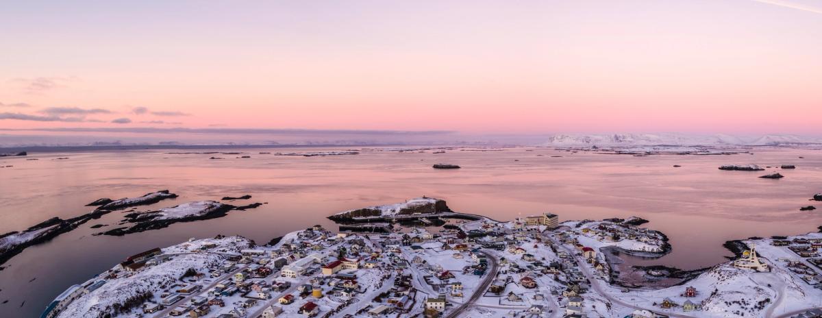 Aerial view of a snow-covered coastal town and rocky islands on a bay, under a pink and purple sunset sky, with ships and distant mountains.