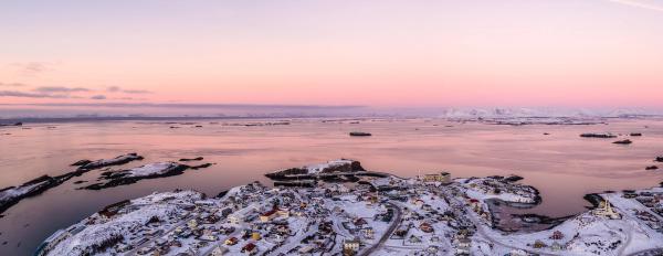 Aerial view of a snow-covered coastal town and rocky islands on a bay, under a pink and purple sunset sky, with ships and distant mountains.