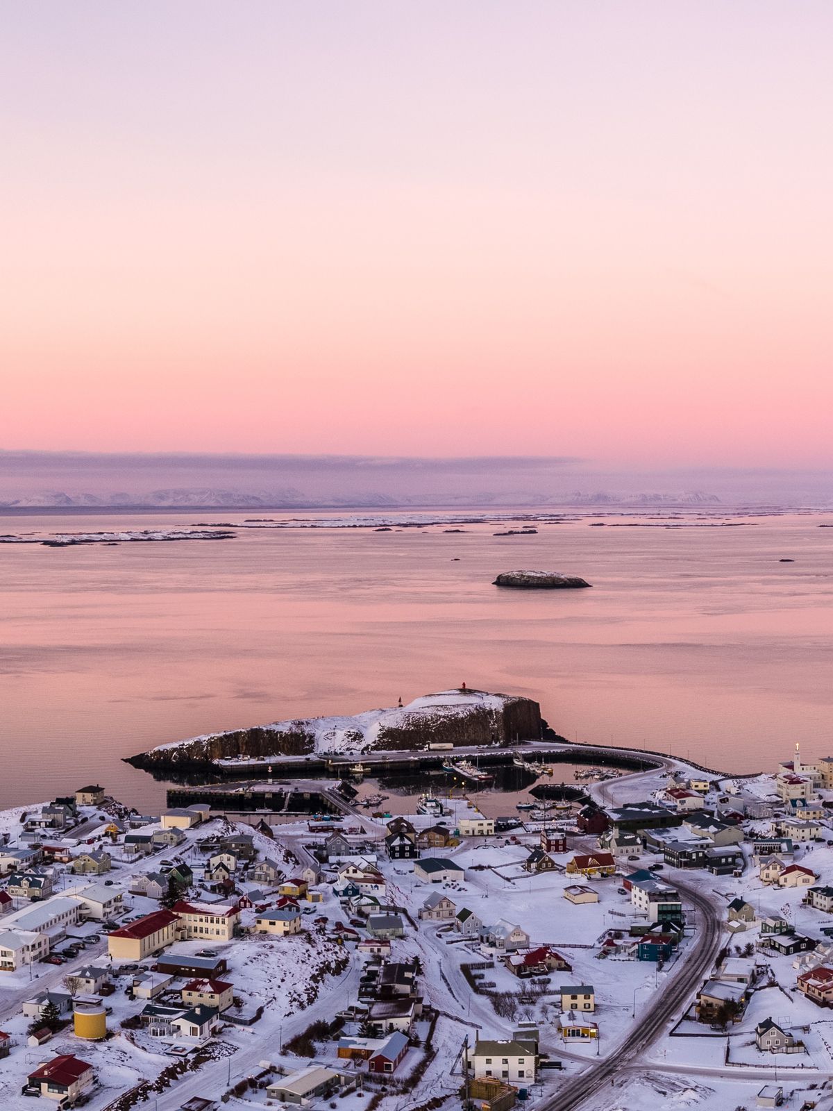 Aerial view of a snow-covered coastal town and rocky islands on a bay, under a pink and purple sunset sky, with ships and distant mountains.