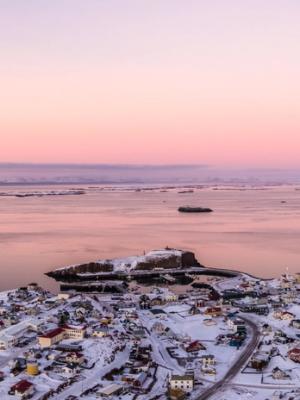 Vista aérea de un pueblo costero cubierto de nieve y de islas rocosas en una bahía, bajo un cielo de atardecer en tonos rosa y morado, con barcos y montañas lejanas al fondo.