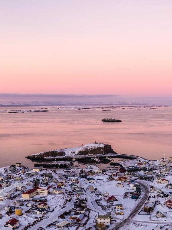 Aerial view of a snow-covered coastal town and rocky islands on a bay, under a pink and purple sunset sky, with ships and distant mountains.