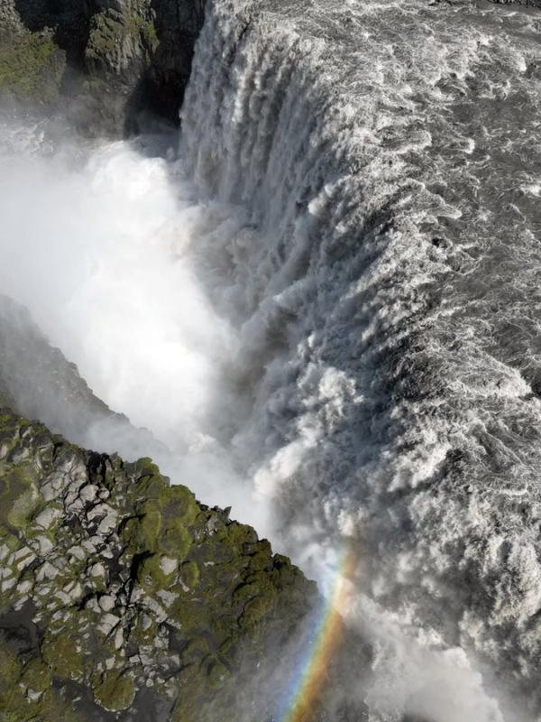 A powerful waterfall plunges into mist, with a rainbow at its base and a line of people visible on a rocky cliff edge to the left.