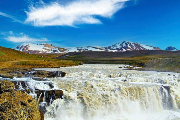A powerful waterfall with churning brown water in a green landscape, backed by snow-capped mountains under a bright blue sky.