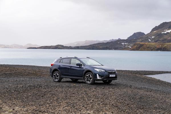 Dark blue Subaru Crosstrek on a gravel beach next to a lake with rocky mountains in the background.