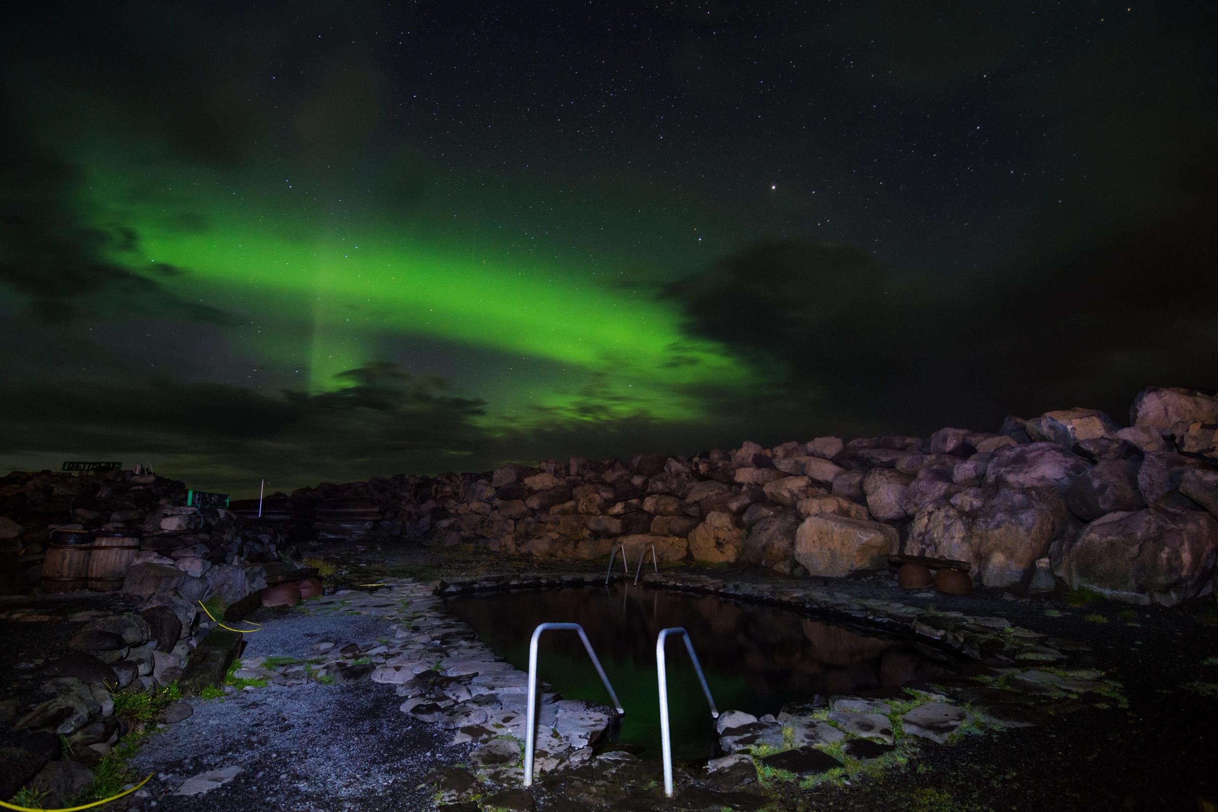 Una piscina de aguas termales bajo una aurora boreal de un verde intenso y un cielo nocturno estrellado.