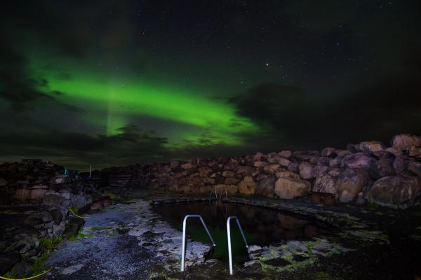 A hot spring pool under a vibrant green aurora borealis and starry night sky.