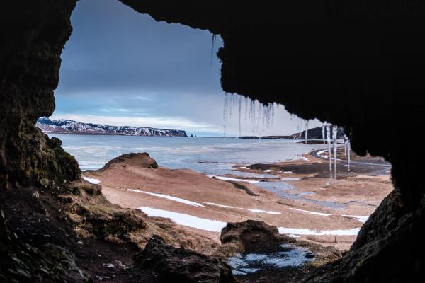 Winter coastal landscape viewed from inside a cave with icicles, showing an icy bay and distant snowy mountains.