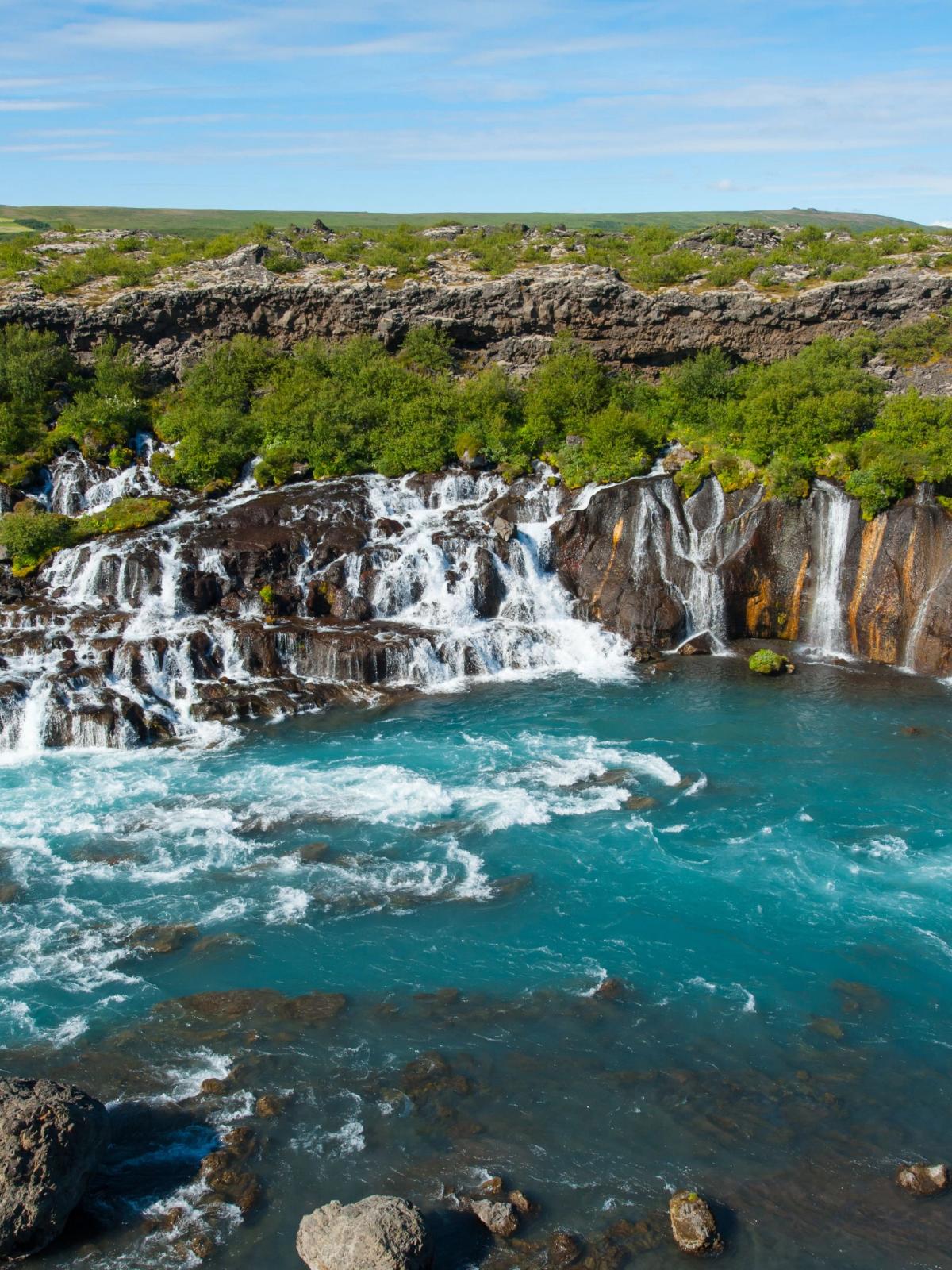 Multiple waterfalls cascade from a rocky, vegetated bank into a vivid turquoise river under a blue sky.