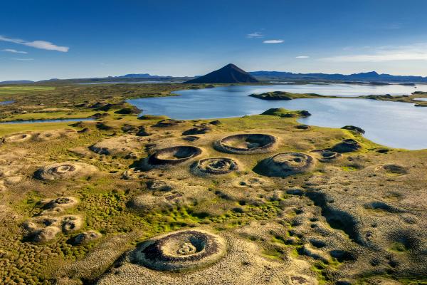 Aerial view of a green landscape with many circular craters, a lake, and a conical mountain.