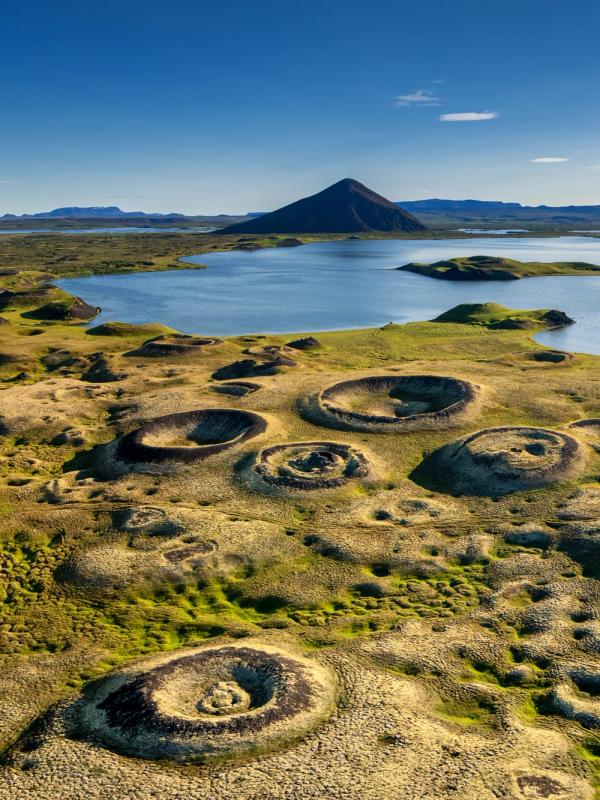 Volcanic landscape with circular, grass-covered craters, a blue lake, and a dark conical mountain under a clear sky.