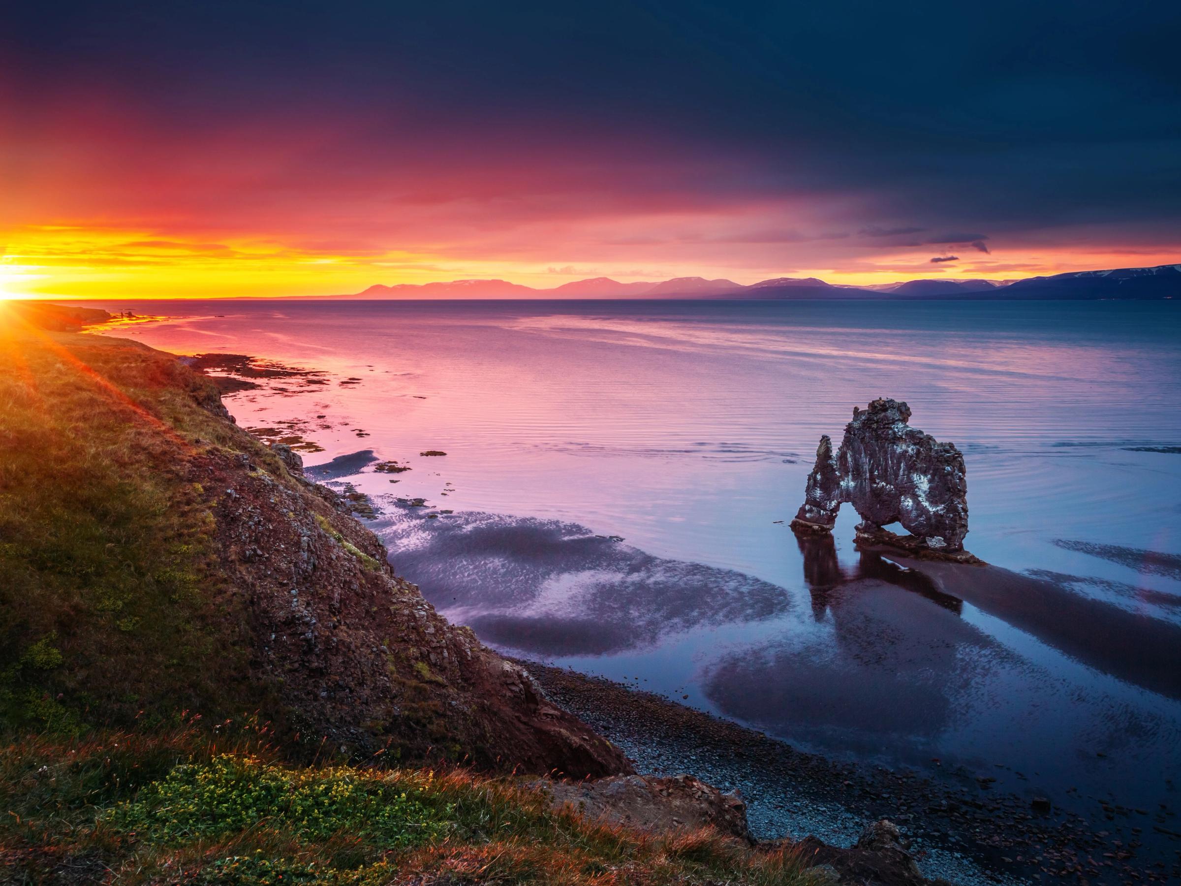 Dramatic sunset over a coastline with an arch-shaped sea stack in calm water, reflecting the colorful sky.