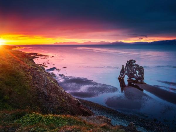 Dramatic sunset over a coastline with an arch-shaped sea stack in calm water, reflecting the colorful sky.