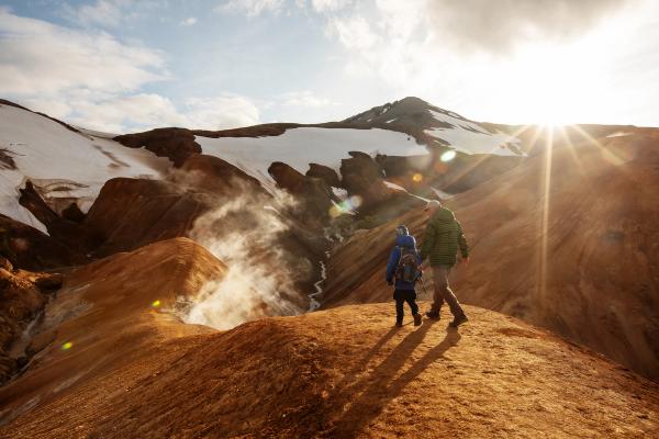 Una familia camina por una cresta rojiza con vistas a un valle geotérmico humeante, montañas nevadas y un sol brillante.