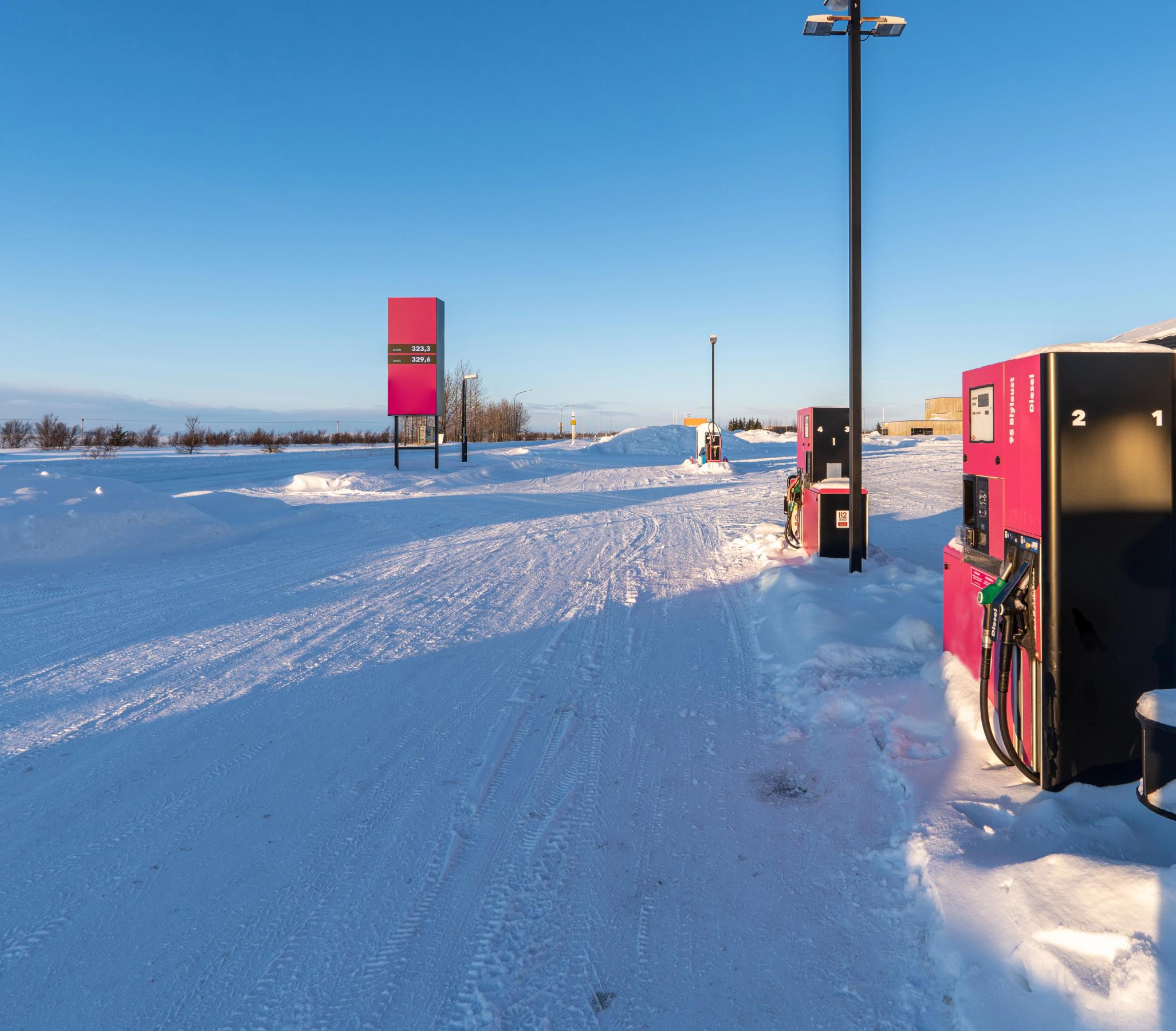 A gas station with pink fuel pumps and a price sign in a snow-covered landscape under a clear blue sky.