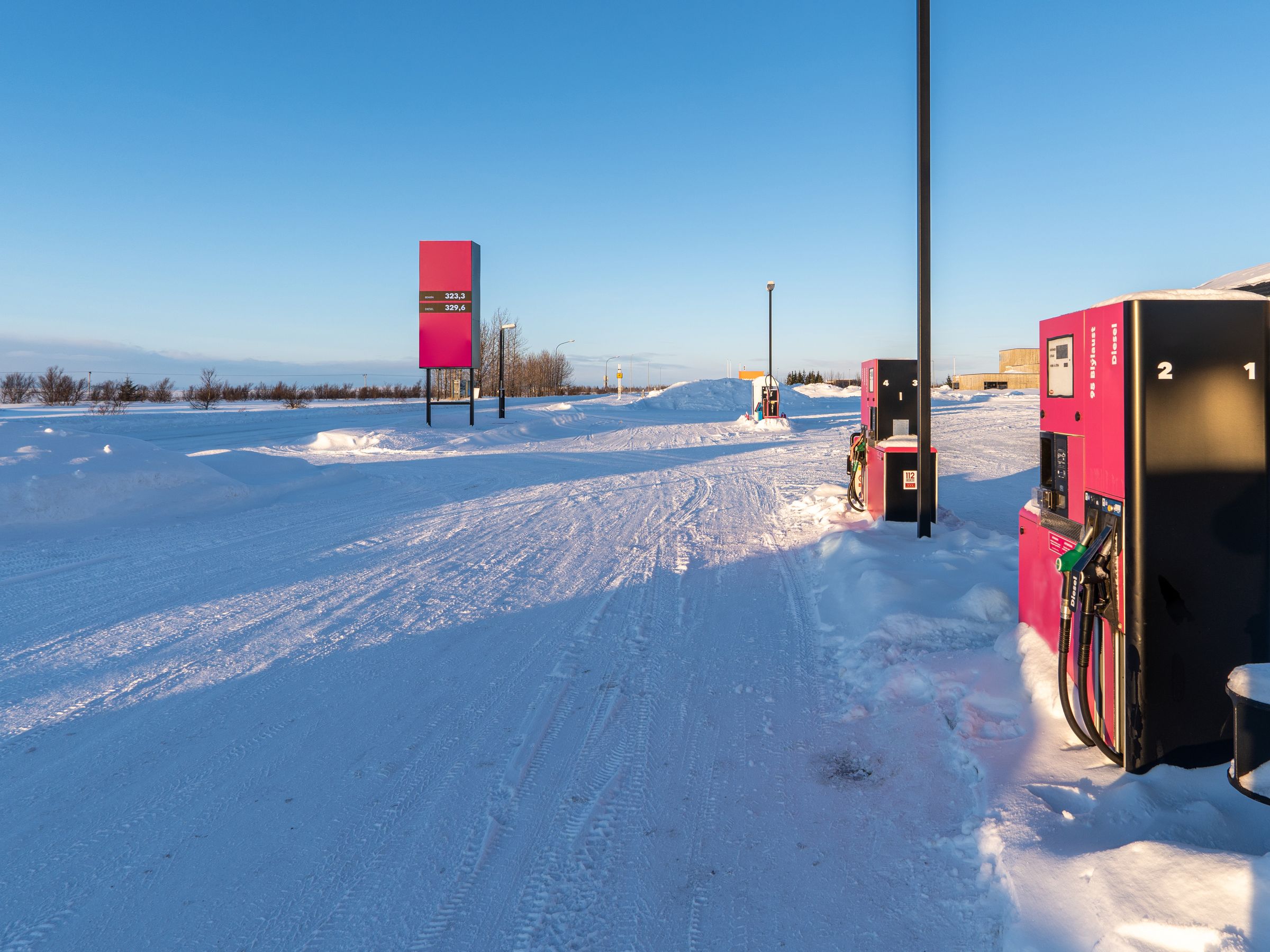 A gas station with pink fuel pumps and a price sign in a snow-covered landscape under a clear blue sky.