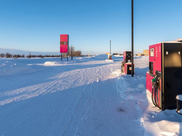 Una gasolinera con surtidores rosas y un cartel de precios en un paisaje nevado bajo un cielo azul despejado.