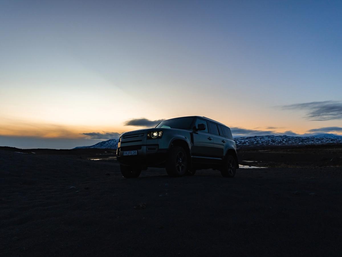 Land Rover Defender al atardecer Land Rover Defender de alquiler aparcado sobre arena negra con los faros encendidos al atardecer en Islandia, disponible en Go Car Rental