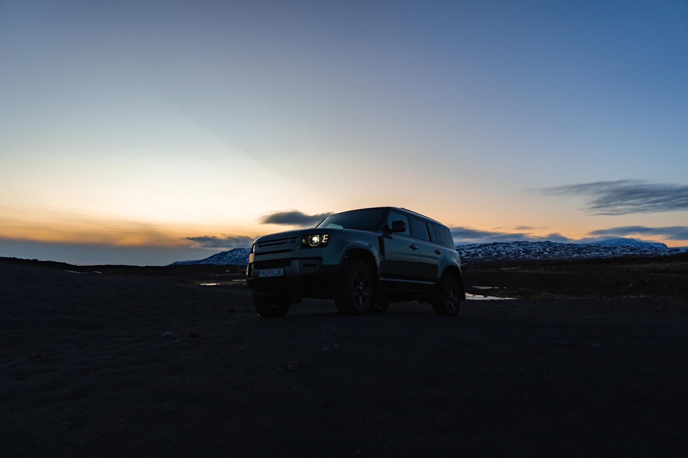 Land Rover Defender al atardecer Land Rover Defender de alquiler aparcado sobre arena negra con los faros encendidos al atardecer en Islandia, disponible en Go Car Rental