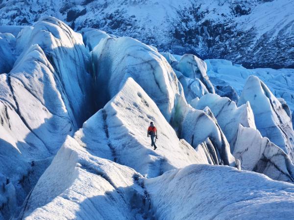 A person in a red jacket walks across a vast, rugged glacier with blue ice formations.