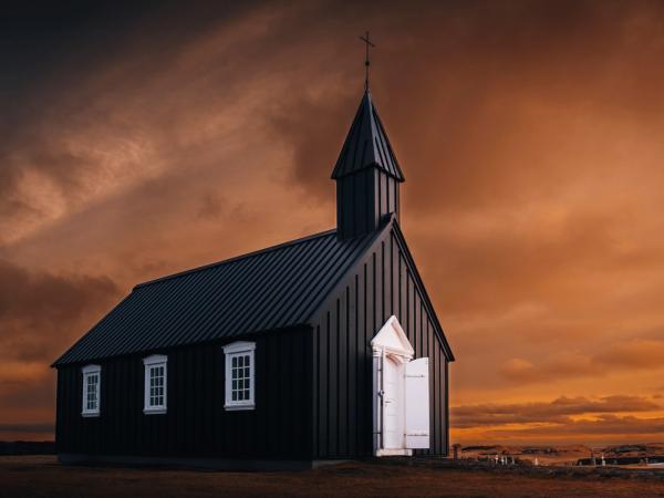 A black church with white windows and an open white door, under a dramatic orange and dark sky.
