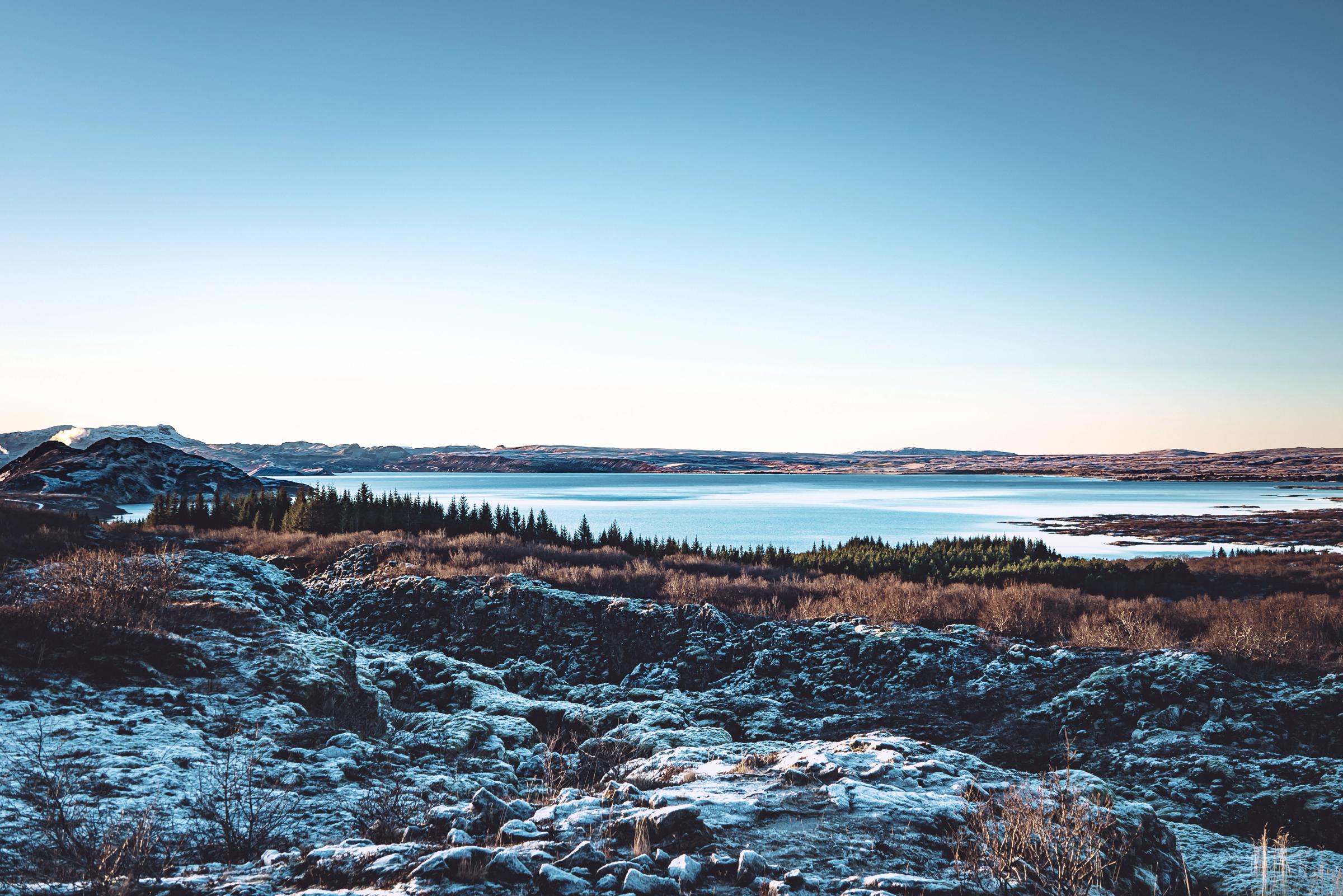 A wide view of a winter landscape with snow-dusted rocks, pine trees, and a tranquil lake under a clear sky.