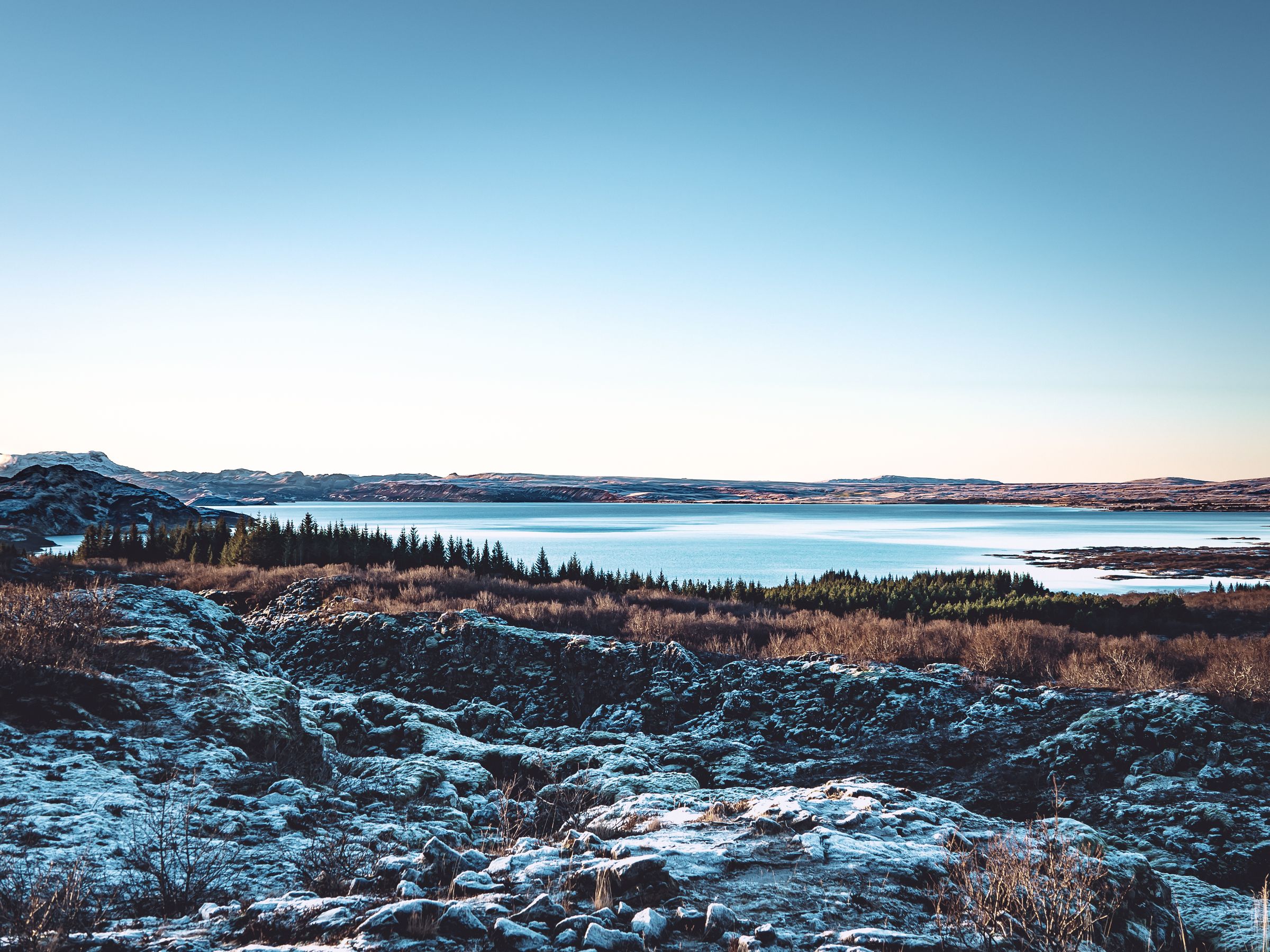 A wide view of a winter landscape with snow-dusted rocks, pine trees, and a tranquil lake under a clear sky.