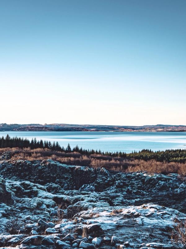 Snow-dusted rocky foreground with sparse vegetation, a blue lake, evergreen trees, and distant hills under a clear sky.