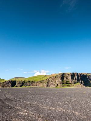 A wide black volcanic plain with tire tracks leads to a long, green-topped mesa under a bright blue sky.